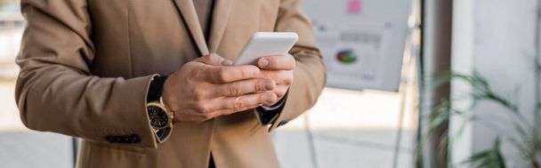 cropped view of accomplished entrepreneur wearing beige blazer and stylish wristwatch, using mobile phone near flip chart with business analytics on blurred background, banner - Фото, зображення