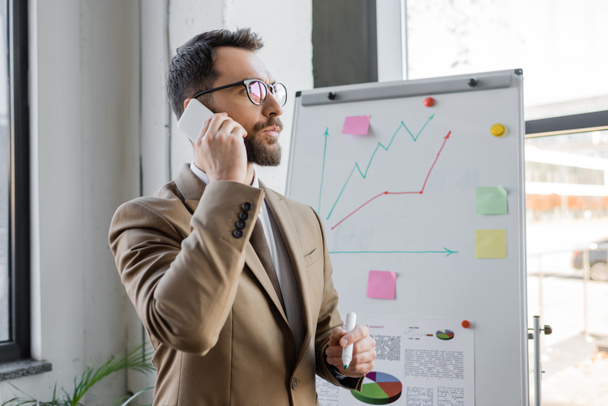 Przemyślany menedżer w stylowym stroju biznesowym i okularach stojących z markerem w pobliżu analityki i wykresów na flip chart i rozmowy przez telefon komórkowy w biurze - Zdjęcie, obraz