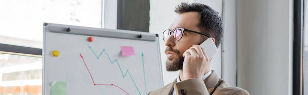 portrait of thoughtful bearded businessman in stylish eyeglasses talking on mobile phone and looking away near flip chart with analytics in office, banner - Photo, Image