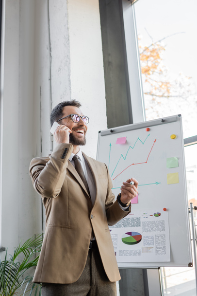 Радостный бородатый менеджер в бежевом пиджаке, галстуке и очках, стоящий с маркером и разговаривающий на мобильном телефоне возле флип-чарта с бизнес-аналитикой и графиками в офисе - Фото, изображение