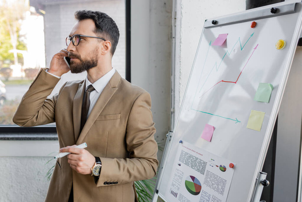 thoughtful bearded businessman in beige blazer and eyeglasses holding marker and looking away while talking on smartphone near market research on flip chart in office - Photo, Image
