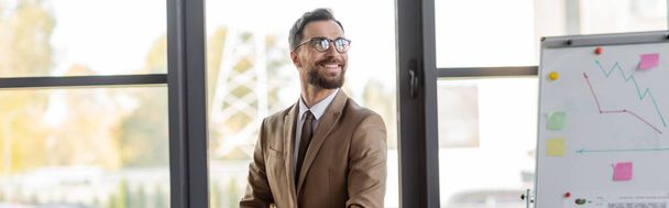 успешный бородатый бизнесмен в стильных очках, бежевый пиджак и галстук глядя в сторону и улыбаясь рядом флип-чарт с графиками и липкие заметки в офисе с большими окнами, баннер - Фото, изображение