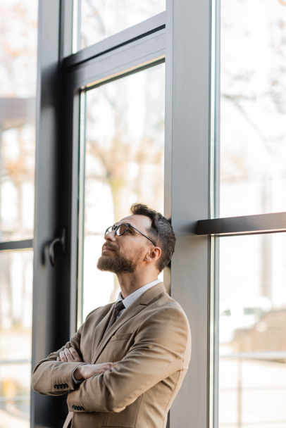 модный и мечтательный бизнесмен в бежевом пиджаке и стильных очках стоит со сложенными руками возле больших окон и смотрит вверх в современном офисе - Фото, изображение