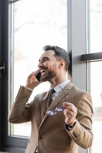 emprendedor alegre y exitoso en blazer beige de moda y corbata hablando en el teléfono móvil y sosteniendo gafas borrosas cerca de ventanas en la oficina moderna - Foto, imagen