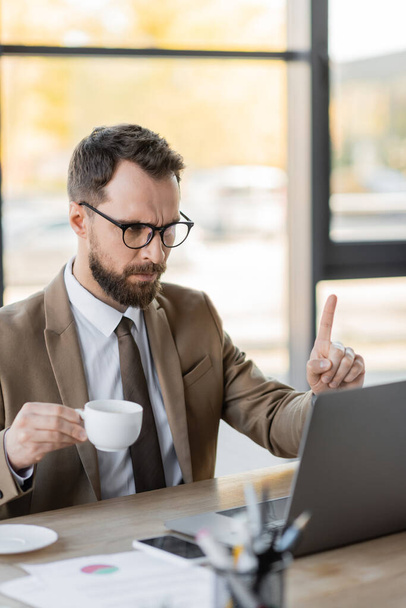 rygorystyczny brodaty menedżer korporacyjny w stylowym stroju biznesowym trzymający filiżankę kawy i pokazujący gest uwagi podczas wideokonferencji na laptopie w pobliżu telefonu komórkowego i dokumentów na biurku - Zdjęcie, obraz