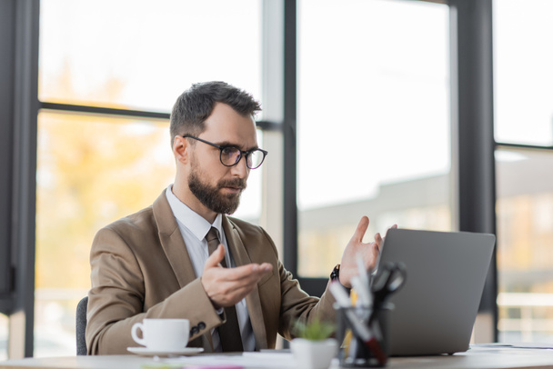 bearded businessman in eyeglasses, stylish blazer and tie gesturing during video conference on laptop near coffee cup and blurred stationery in office - Foto, imagen