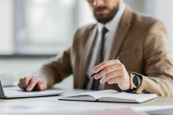 частковий вигляд корпоративного менеджера в бежевому блістері, краватці та розкішному наручному годиннику, що сидить з ручкою біля ноутбука під час роботи на ноутбуці на робочому місці в офісі
 - Фото, зображення