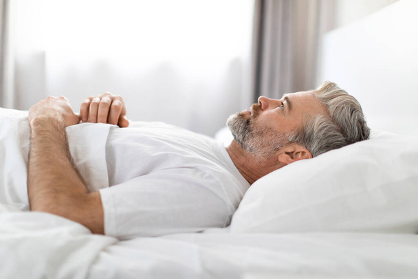 Droevige knappe man van middelbare leeftijd met een baard en een wit t-shirt onder een deken in bed, kijkend naar het plafond, hebben zware gedachten na het opstaan in de ochtend, zijaanzicht - Foto, afbeelding