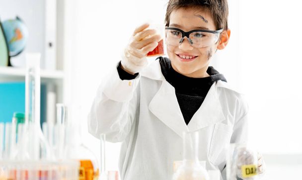 School boy with dirty face wearing protection glasses looking at liquid in glass during chemistry experiment in elementary science class and smiling. Clever pupil in lab during test - Photo, image