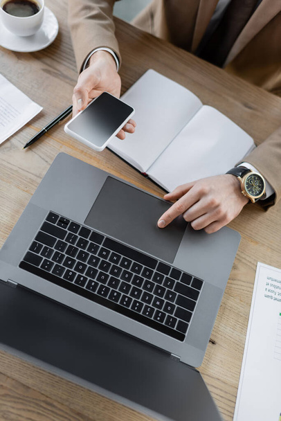 widok z góry przycięty biznesmen w luksusowym zegarku na rękę za pomocą touchpad laptopa, trzymając smartfona z pustym ekranem w pobliżu pustego notebooka, długopisu, dokumentów i filiżanki kawy na stole - Zdjęcie, obraz