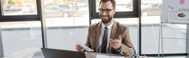 ευτυχής και επιτυχημένος επιχειρηματίας με κομψά γυαλιά, μπεζ σακάκι και γραβάτα κρατώντας έγγραφο και δείχνοντας με το χέρι κατά τη διάρκεια τηλεδιάσκεψης στο φορητό υπολογιστή κοντά σημειωματάρια στο σύγχρονο γραφείο, πανό - Φωτογραφία, εικόνα