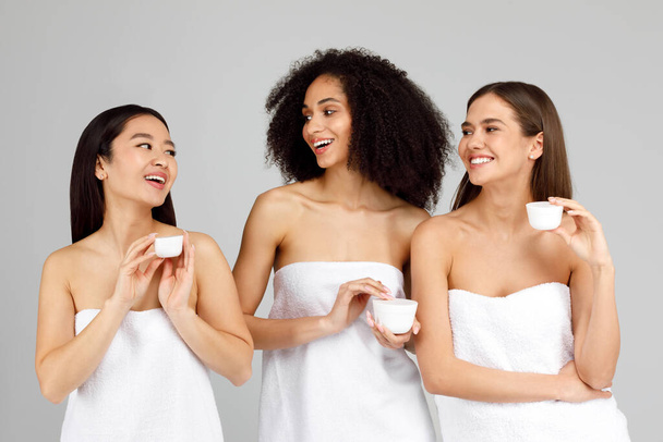 Τρεις νεαρές πολύμορφες γυναίκες που κατέχουν βάζα καλλυντικής φυσικής κρέμας προσώπου, πολυφυλετικές κυρίες που φροντίζουν το δέρμα τους, υποβάλλονται σε αντιγηραντικές θεραπείες, απολαμβάνοντας την ημέρα spa - Φωτογραφία, εικόνα