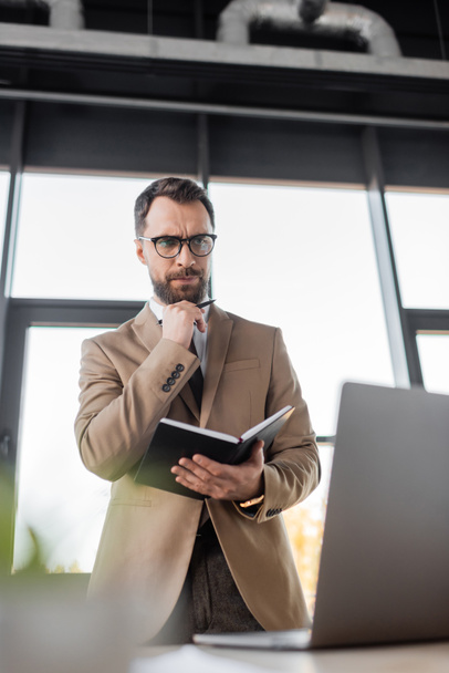 low angle view of thoughtful businessman in eyeglasses and stylish blazer standing with notebook and pen and looking at laptop in office on blurred foreground - Photo, Image