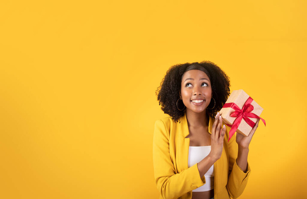 Ideas for gift. Excited glad black woman holding gift box and looking at blank space isolated on yellow background, studio shot. Woman thinking of anniversary, Christmas and New Year present - Photo, Image