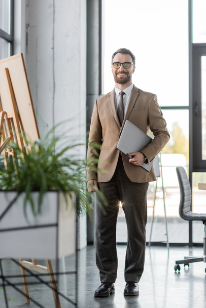 full length on successful bearded businessman in trendy blazer, pants, tie and eyeglasses holding laptop and smiling at camera near flip chart and blurred plants in modern office - Фото, изображение