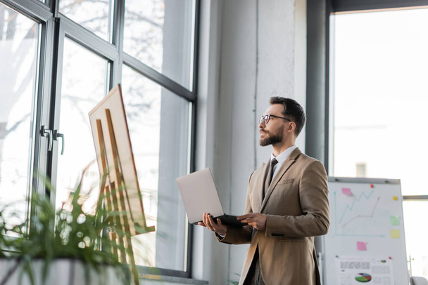confident and fashionable bearded businessman with laptop looking away through windows near flip charts with infographics and blurred plants in contemporary office - Photo, Image