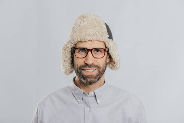 Headshot de chico caucásico guapo alegre lleva una cálida gorra de invierno con orejeras, gafas y camisa blanca, mira directamente a la cámara, listo para pasear al aire libre, aislado sobre fondo blanco
 - Foto, imagen