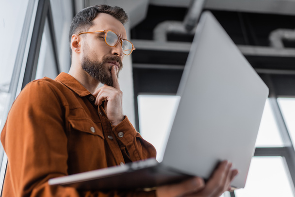 low angle view of serious bearded businessman in trendy eyeglasses and shirt looking at laptop and thinking while standing in office on blurred foreground, corporate lifestyle  - Foto, imagen