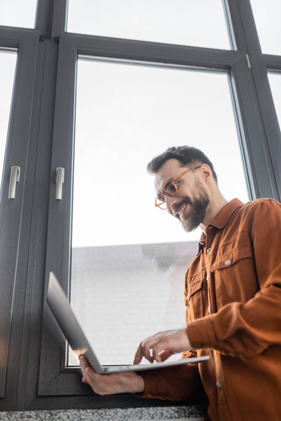 vue à angle bas de l'homme d'affaires réussi et barbu dans les lunettes et chemise élégante debout près de la fenêtre dans le bureau moderne et souriant tout en utilisant un ordinateur portable, mode de vie d'entreprise  - Photo, image