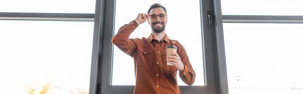low angle view of cheerful and accomplished businessman in stylish casual clothes holding coffee to go, adjusting eyeglasses and smiling at camera in office, banner - Photo, Image