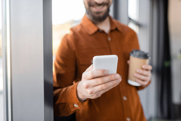częściowy widok brodatego biznesmena w stylowej koszuli wiadomości na telefon komórkowy, stojąc z kawą w jednorazowym kubku w biurze, rozmyte tło - Zdjęcie, obraz