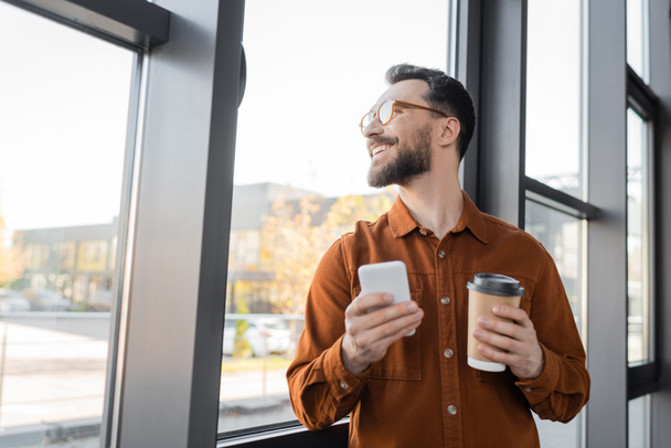 χαρούμενη, επιτυχημένη και γενειοφόρος επιχειρηματίας σε κομψό πουκάμισο και γυαλιά ηλίου στέκεται με smartphone και takeaway ποτό κοντά στο παράθυρο στο γραφείο και κοιτάζοντας μακριά, εταιρικό τρόπο ζωής  - Φωτογραφία, εικόνα