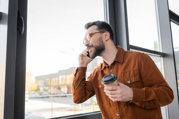ευχαριστημένος γενειοφόρος επιχειρηματίας σε μοντέρνα γυαλιά ηλίου και κομψό πουκάμισο κρατώντας χάρτινο κύπελλο κατά τη διάρκεια της συνομιλίας στο smartphone κοντά στο παράθυρο του σύγχρονου γραφείου - Φωτογραφία, εικόνα