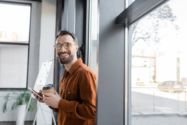 красивый и радостный бизнесмен в стильной рубашке и очках стоит с одноразовой чашкой и мобильным телефоном рядом с окнами в офисе и смотрит в камеру - Фото, изображение