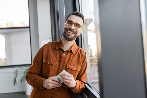 опытный и харизматичный бизнесмен в стильных очках и рубашке, стоящий с мобильным телефоном возле окна и улыбающийся на камеру в офисе, размытый передний план - Фото, изображение