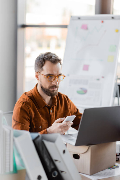fokussierter bärtiger Geschäftsmann in trendiger Brille und Hemd sitzt mit Handy neben Ordnern, Laptop auf Karton und Flipchart auf verschwommenem Hintergrund im Büro - Foto, Bild