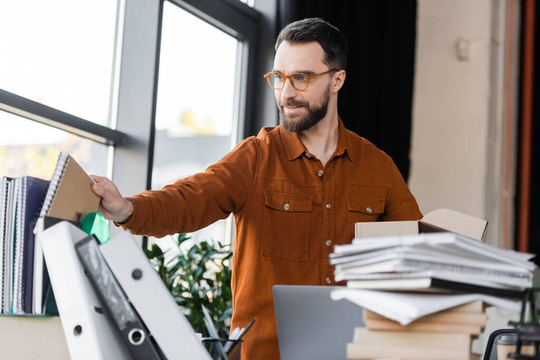 позитивный бородатый бизнесмен в рубашке и очках с коробкой в руках и с ноутбуком на рабочем месте с ноутбуком, много ноутбуков и папок в офисе - Фото, изображение