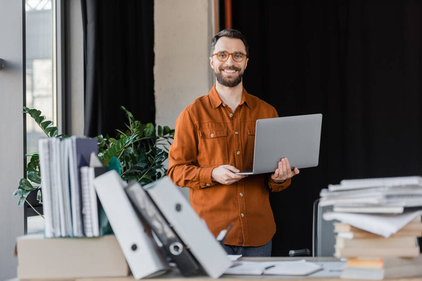 happy successful businessman in stylish eyeglasses and shirt standing with laptop and looking at camera near pile of books, notebooks and folders on blurred foreground in office - Zdjęcie, obraz