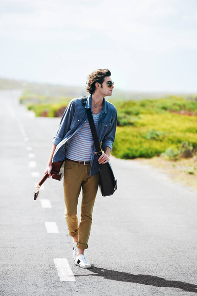 Getting some musical inspiration on the open road. Trendy young male wearing sunglasses walking down the middle of a road holding a guitar - Fotografie, Obrázek