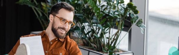 brunette bearded businessman in fashionable eyeglasses and shirt smiling near document and green decorative plants in contemporary office on blurred background, banner - Photo, Image