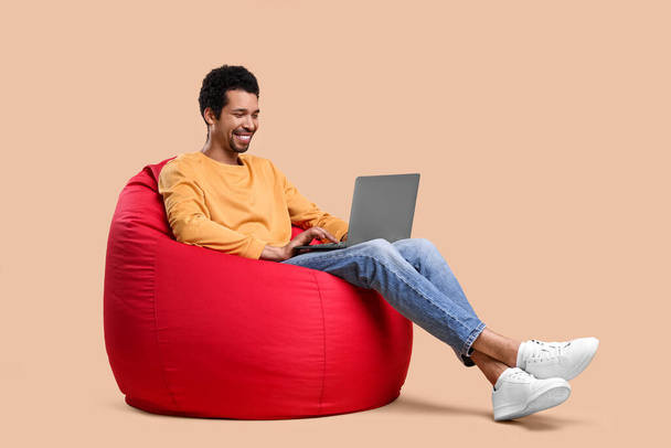 Ευτυχισμένος άνθρωπος με φορητό υπολογιστή κάθεται σε καρέκλα beanbag σε μπεζ φόντο - Φωτογραφία, εικόνα