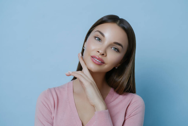 Beautiful dark haired woman with applied makeup, healthy glowing skin, touches cheek gently, wears rosy jumper, looks directly at camera, isolated over blue background. Women and beauty concept - Foto, imagen