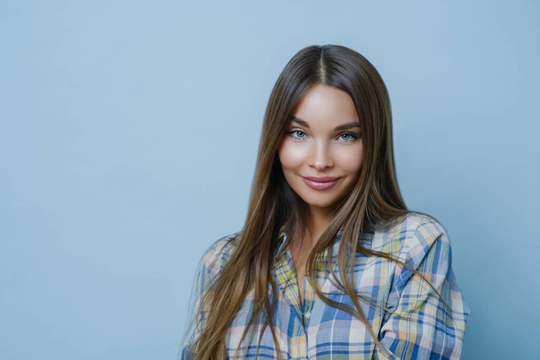 Portrait of good looking female student poses against blue studio wall, looks at camera with charming smile, dresses checkered shirt, poses for university photo, feels optimistic, wears make up - Photo, Image