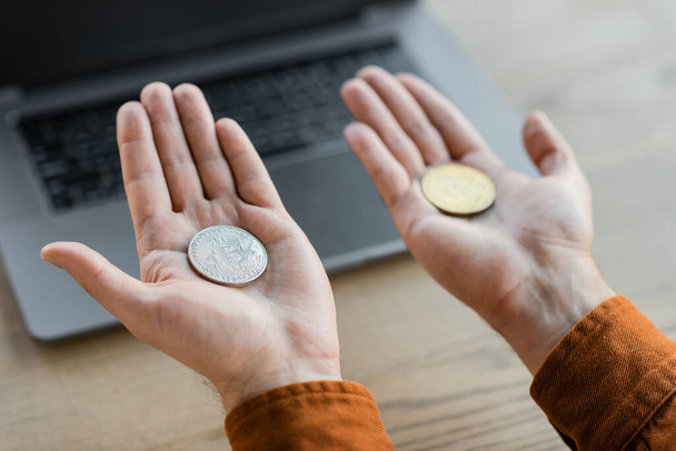 KYIV, UKRAINE - OCTOBER 18, 2022: partial view of successful businessman holding golden and silver bitcoins on open hand palms near blurred laptop on work desk in office - Photo, image