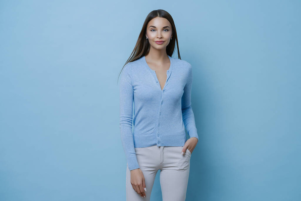 Cute enthusiastic woman has slim figure, wears casual jumper and white jeans, keeps hand in pocket, stands confident, walks with friends, has pleasant conversation, poses against blue background - Valokuva, kuva