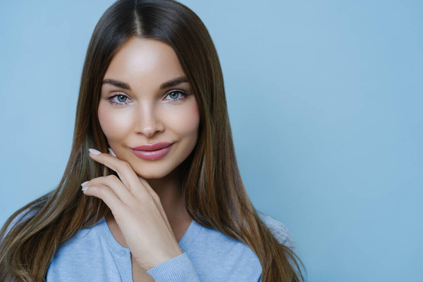 Natural magnetic young woman with fresh clean skin, has attractive facial features, gazes sensually at camera, feels beautiful and appealing, touches jawline, dressed in casual jumper isolated on blue - Fotografie, Obrázek