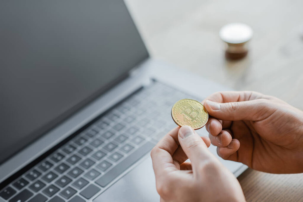 KYIV, UKRAINE - OCTOBER 18, 2022: cropped view of ambitious businessman holding golden bitcoin near laptop with blank screen on office desk, blurred background - Fotoğraf, Görsel