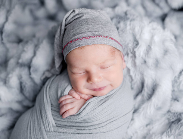 Cute newborn baby boy swaddled in grey fabric sleeping on fur blanket. Adorable infant child kid napping studio portrait - Фото, зображення