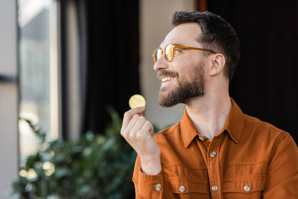 KYIV, UKRAINE - OCTOBER 18, 2022: joyful and accomplished businessman in stylish eyeglasses and shirt holding golden bitcoin, smiling and looking away in modern office on blurred background - Fotó, kép