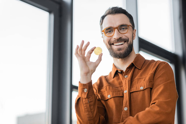 KYIV, UKRAINE - OCTOBER 18, 2022: bearded businessman in fashionable shirt and eyeglasses holding golden bitcoin and smiling at camera near blurred windows in modern office on blurred background - Photo, Image
