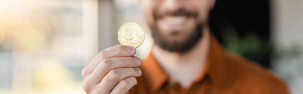 KYIV, UKRAINE - OCTOBER 18, 2022: selective focus of golden bitcoin in hand of cropped bearded businessman smiling on blurred background in office, banner - 写真・画像