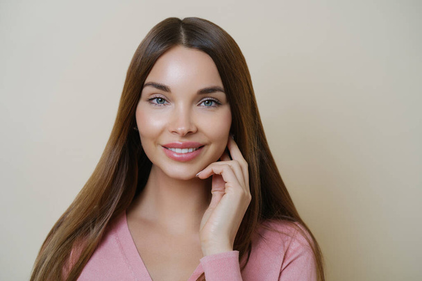 Adorable young female model smiles pleasantly, has long dark hair, looks directly at camera with satisfied expression, has perfect clean skin, wears rosy jumper, isolated over beige background - Zdjęcie, obraz