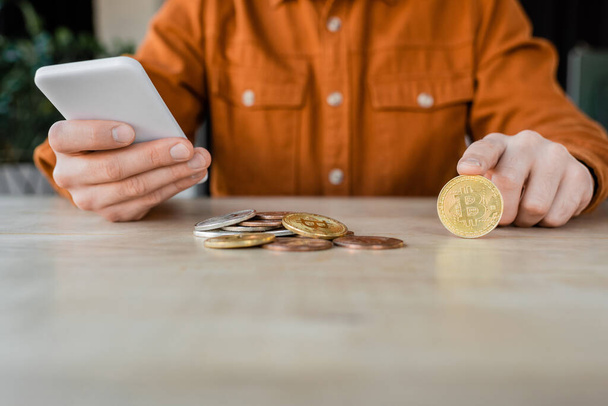 KYIV, UKRAINE - OCTOBER 18, 2022: partial view of entrepreneur in stylish shirt holding mobile phone while sitting near silver and golden bitcoins on work desk in office - Foto, Bild