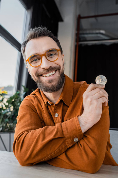 KYIV, UKRAINE - OCTOBER 18, 2022: overjoyed and bearded businessman in fashionable eyeglasses and stylish shirt holding silver bitcoin and smiling at camera while sitting in modern office - Фото, изображение