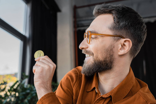 KYIV, UKRAINE - OCTOBER 18, 2022: brunette bearded businessman, pleased and successful, wearing trendy eyeglasses, looking at golden bitcoin in contemporary office on blurred background - 写真・画像