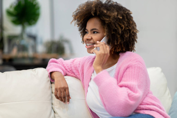 Happy cheerful smiling pretty millennial black woman in comfy casual outwear sitting on couch, having phone conversation with friend or lover at home, looking at copy space for advertisement - 写真・画像